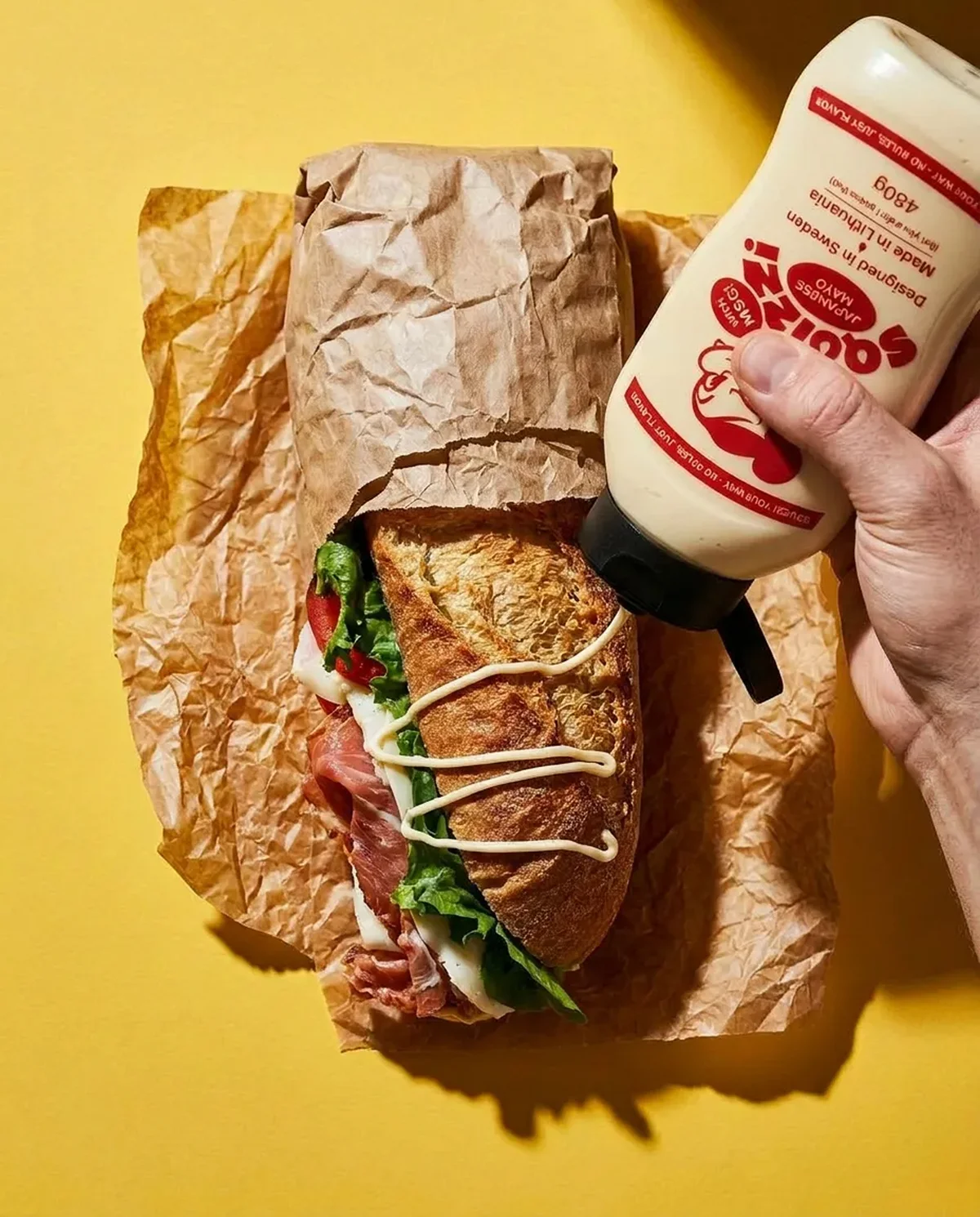 squizzi – Hand drizzling Squizzi mayo onto a baguette over a yellow backdrop