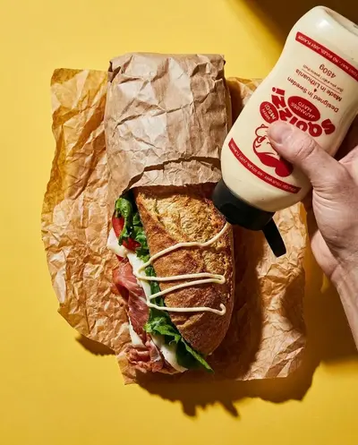 Hand drizzling Squizzi mayo onto a baguette over a yellow backdrop – squizzi